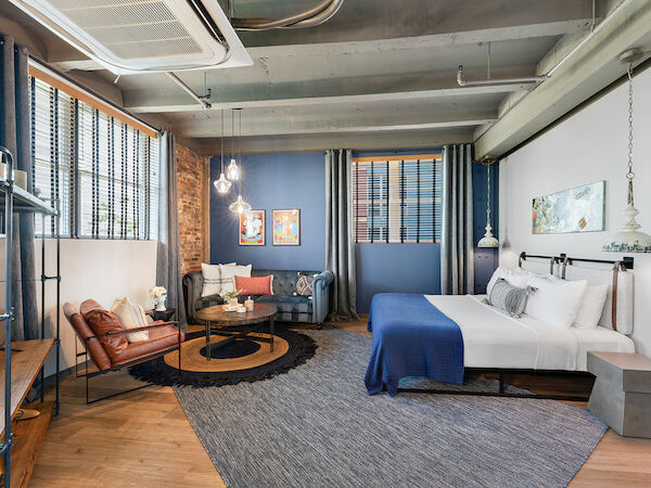 A modern, stylish bedroom with industrial elements, featuring a large bed, sitting area with chairs, coffee table, and blue accents throughout the room.
