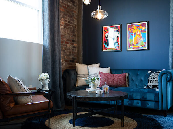 A cozy living room with a brown armchair, blue couch, round coffee table, two framed posters on a blue wall, and decorative plants.