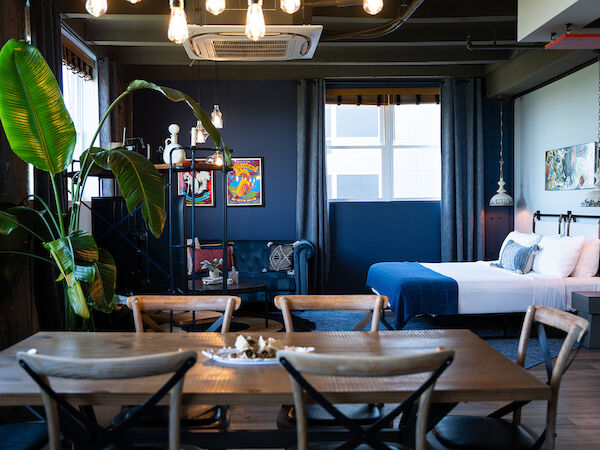 This image shows a modern, stylish room with a bed, dining area, plants, wall art, and natural light from windows.