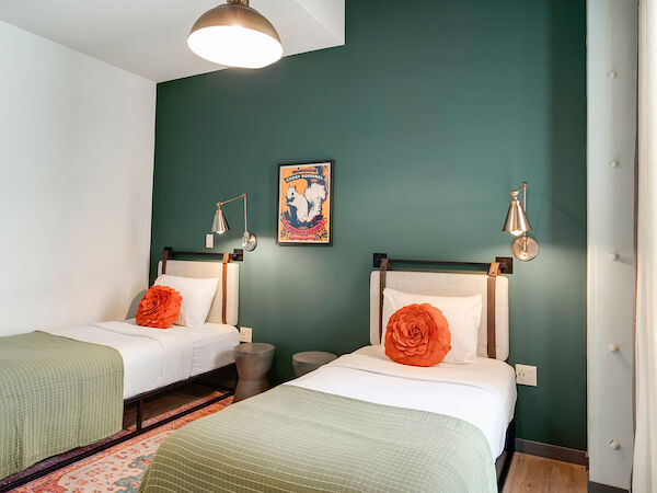The image shows a neatly arranged room with two single beds, green bedding, orange pillows, and a green accent wall adorned with a framed picture.