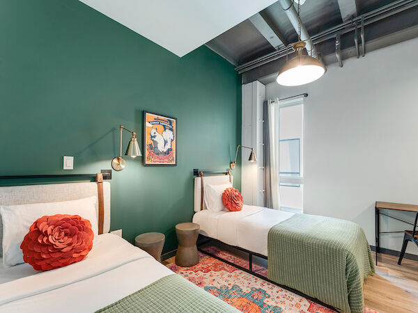 The image shows a well-lit room with two single beds, green accent walls, red pillows, a small desk, and modern décor, creating a cozy ambiance.