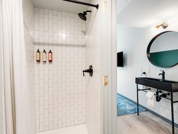 The image shows a modern bathroom with a tiled shower, round mirror, black vanity with a sink, and a wall-mounted TV in the background.