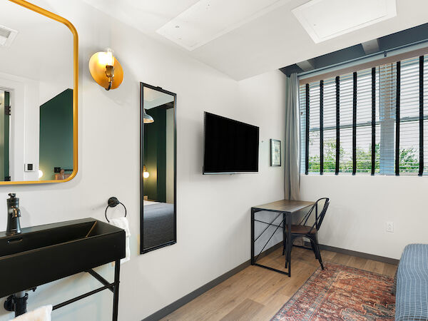 A modern room features a wall-mounted TV, desk with chair, sink, mirror, and window with blinds, accented with a rug and wall lighting.