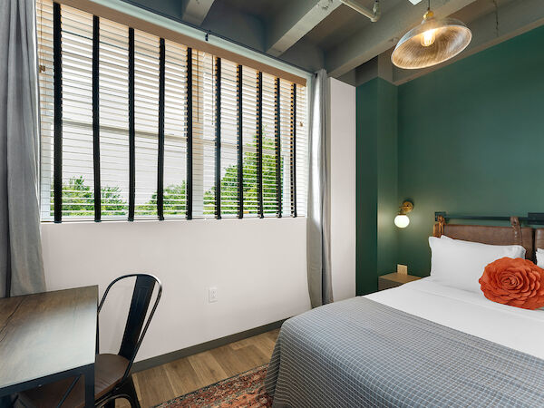 A bedroom with green walls, a desk and chair, bed with gray bedding and a red pillow, large window with blinds, and a ceiling light fixture.