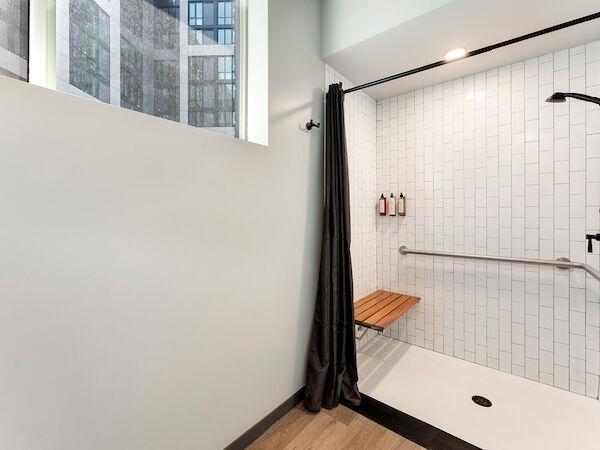 A modern, accessible shower with a bench, grab bar, and adjustable showerhead, featuring white tile walls and a black curtain.