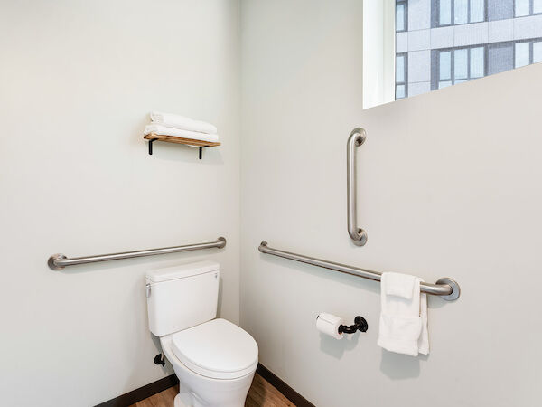 A clean, modern bathroom with a toilet, grab bars, a towel rack with towels, and a window with a view of a building outside.