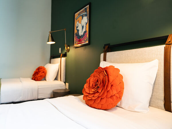 Two beds with white linens and large orange flower pillows, green-painted wall, framed artwork, and a wall lamp in a modern bedroom.
