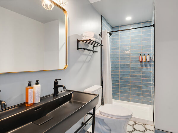 The image shows a modern bathroom with a double sink vanity, a large mirror, a toilet, and a shower with blue tiles.