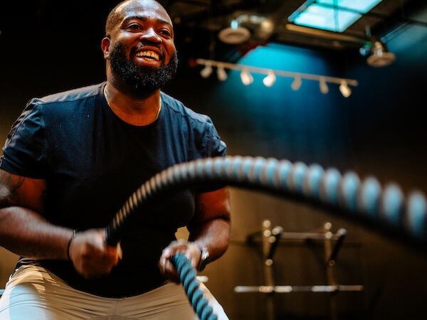 A person is exercising with battle ropes in a gym, smiling and wearing a black shirt and shorts in a well-lit space.
