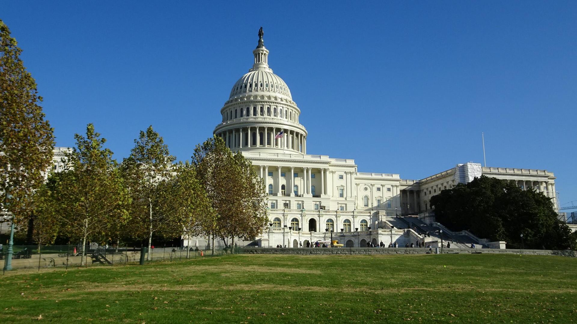 The image shows the United States Capitol building with a clear blue sky and a green lawn in the foreground.