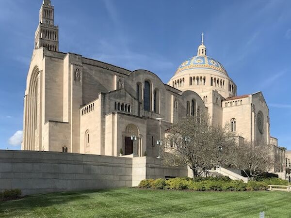 The image shows a large cathedral with a prominent dome and tower, set against a clear blue sky, surrounded by green lawns and trees.
