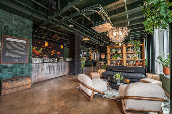 The image shows a stylish lounge with modern furniture, plants, a decorative chandelier, and a bar area.