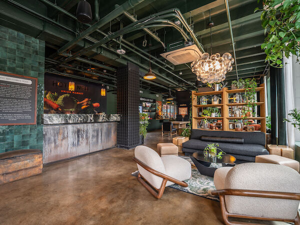 The image shows a stylish lounge with modern furniture, plants, a decorative chandelier, and a bar area.