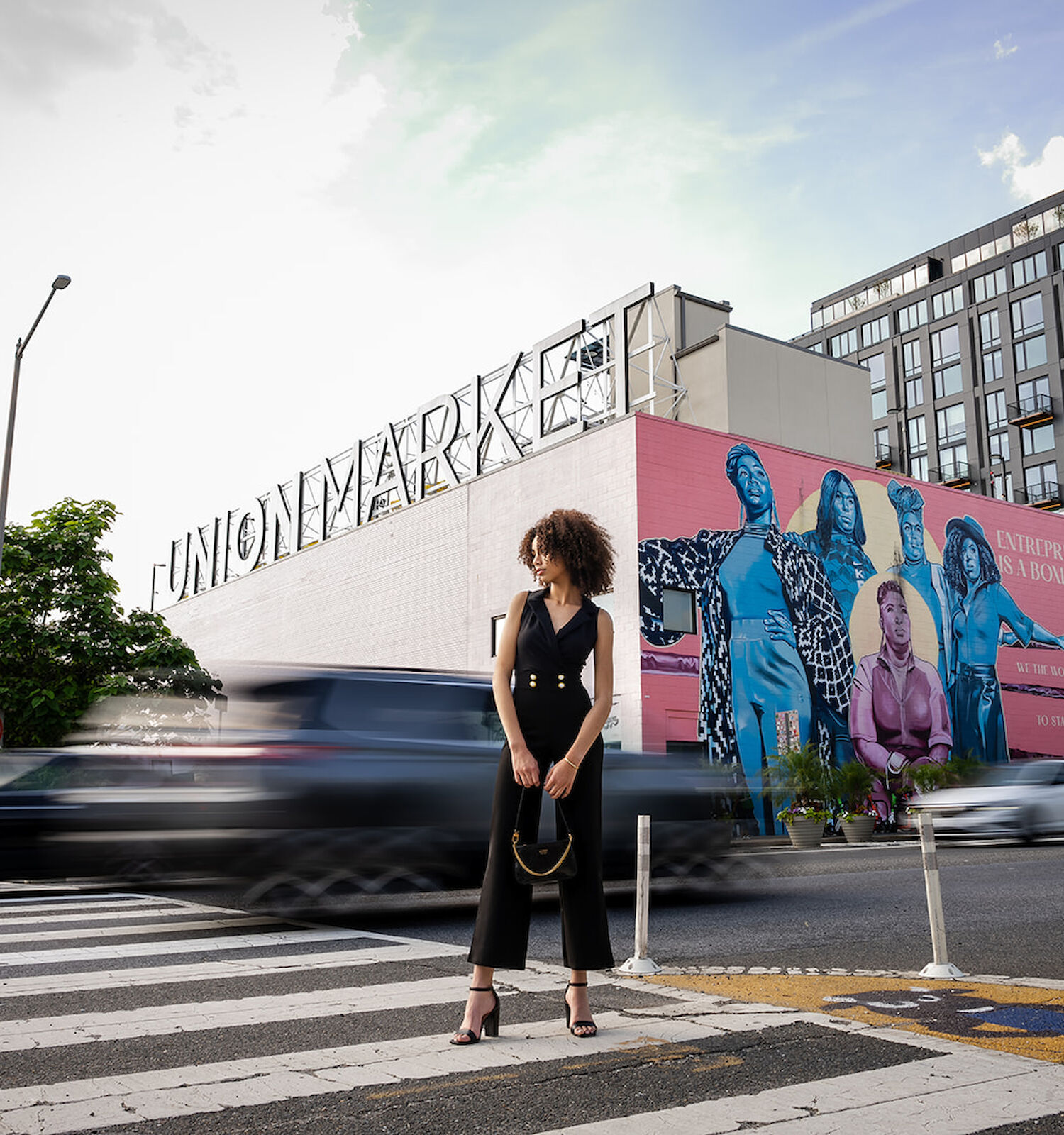 A person stands on a crosswalk in front of a building with a large colorful mural and passing cars, under a partly cloudy sky.