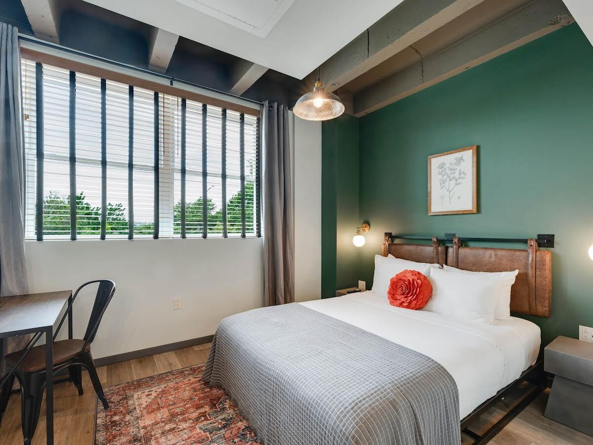 A cozy bedroom with a bed, decorative pillows, pendant lights, a desk, chair, large window, green accent wall, and framed artwork.