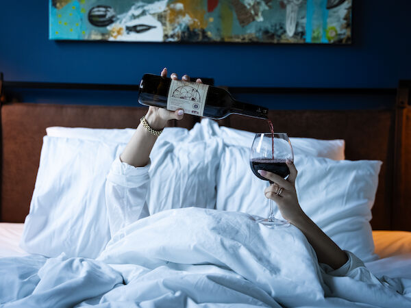 A couple toasting with wine in bed, wearing a VR headset and holding a portable device above them, with blue-lit bedroom ambiance.
