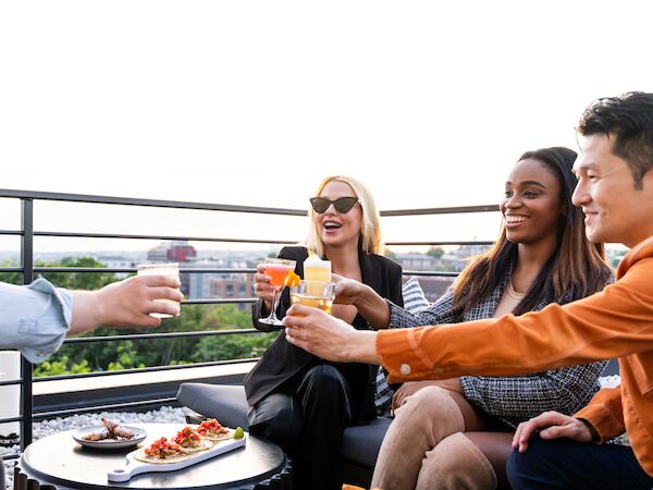 A group of friends on a rooftop toasting drinks, enjoying appetizers and photos with a city view in the background.