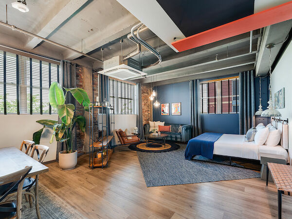 A modern, open-plan living space with a bed, seating area, dining table, and large plants, featuring exposed ceilings and bright decor.