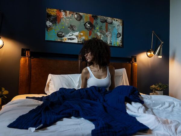 A person sits up in bed under a dark blue wall, white sheets, and a blue blanket, with warm wall lamps and abstract art above the headboard.