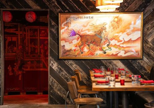 A cozy restaurant with a large oriental painting, wooden tables set with red napkins, and warm lighting.