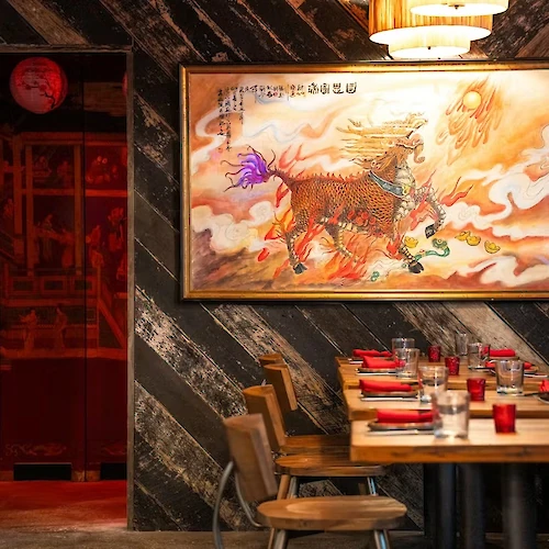 A cozy restaurant with a large oriental painting, wooden tables set with red napkins, and warm lighting.