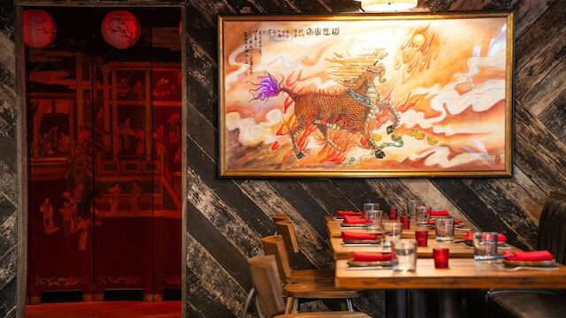 A cozy restaurant with a large oriental painting, wooden tables set with red napkins, and warm lighting.