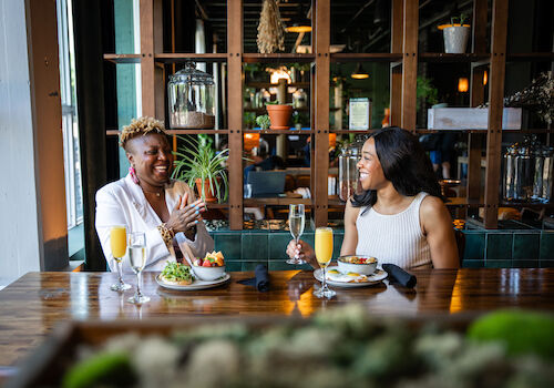 Two people dining at a table with breakfast? drinks like mimosas, salads and plates, in a stylish restaurant setting with plants in the background.
