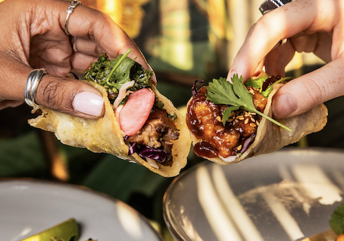 Two hands share bite-sized tacos filled with meat, herbs, and greens, while colorful skewers and plated tacos appear in the background.