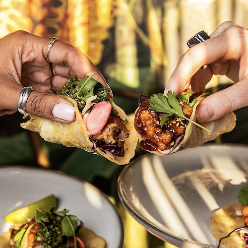 Two hands share bite-sized tacos filled with meat, herbs, and greens, while colorful skewers and plated tacos appear in the background.