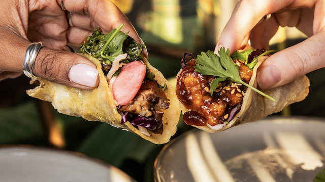 Two hands share bite-sized tacos filled with meat, herbs, and greens, while colorful skewers and plated tacos appear in the background.