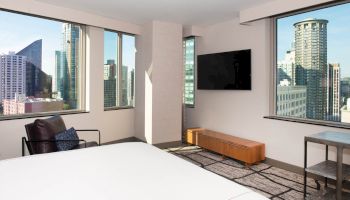 The image shows a modern hotel room with a bed, a chair, a flat-screen TV, and large windows offering a cityscape view.