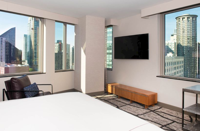 The image shows a modern hotel room with a bed, a chair, a flat-screen TV, and large windows offering a cityscape view.