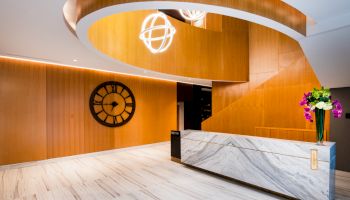 This image shows a modern lobby with a marble reception desk, wooden walls, a large clock, and contemporary lighting fixtures.