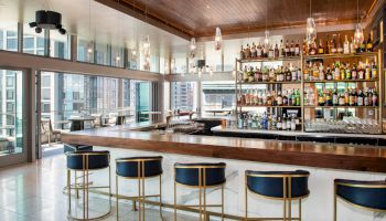 A modern bar with sleek stools, a well-stocked liquor shelf, pendant lights, and large windows offering a city view.