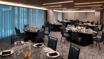 A banquet hall is set up with round tables, black chairs, white and black place settings, floral centerpieces, and evenly spaced lighting fixtures.