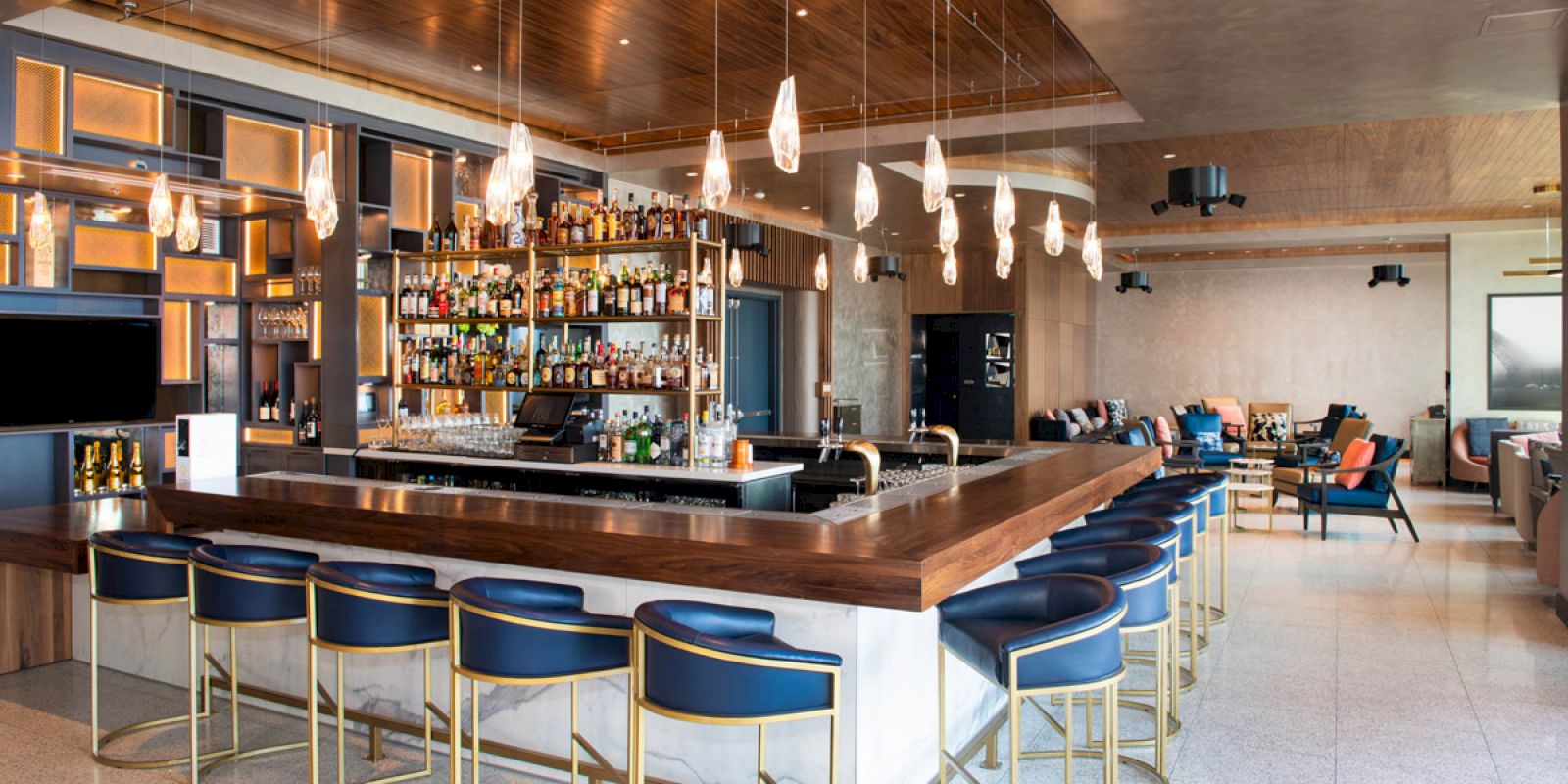 This image shows a modern bar area with stylish seating, a well-stocked bar, pendant lighting, and a lounge area in the background.