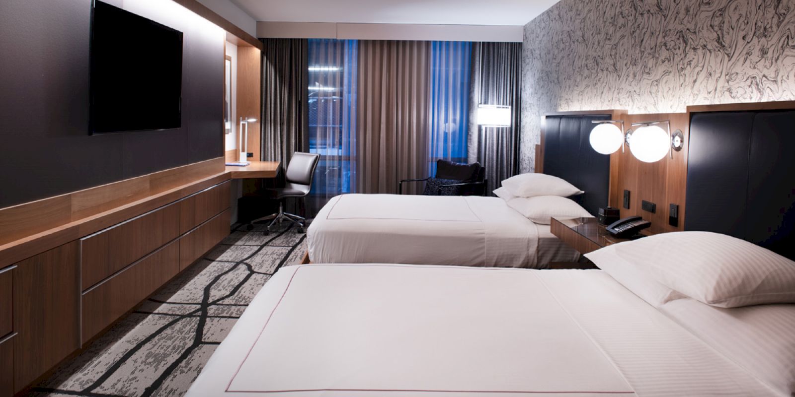 A modern hotel room with two double beds, a TV, a desk with a chair, and floor-to-ceiling curtains covering a window is shown.