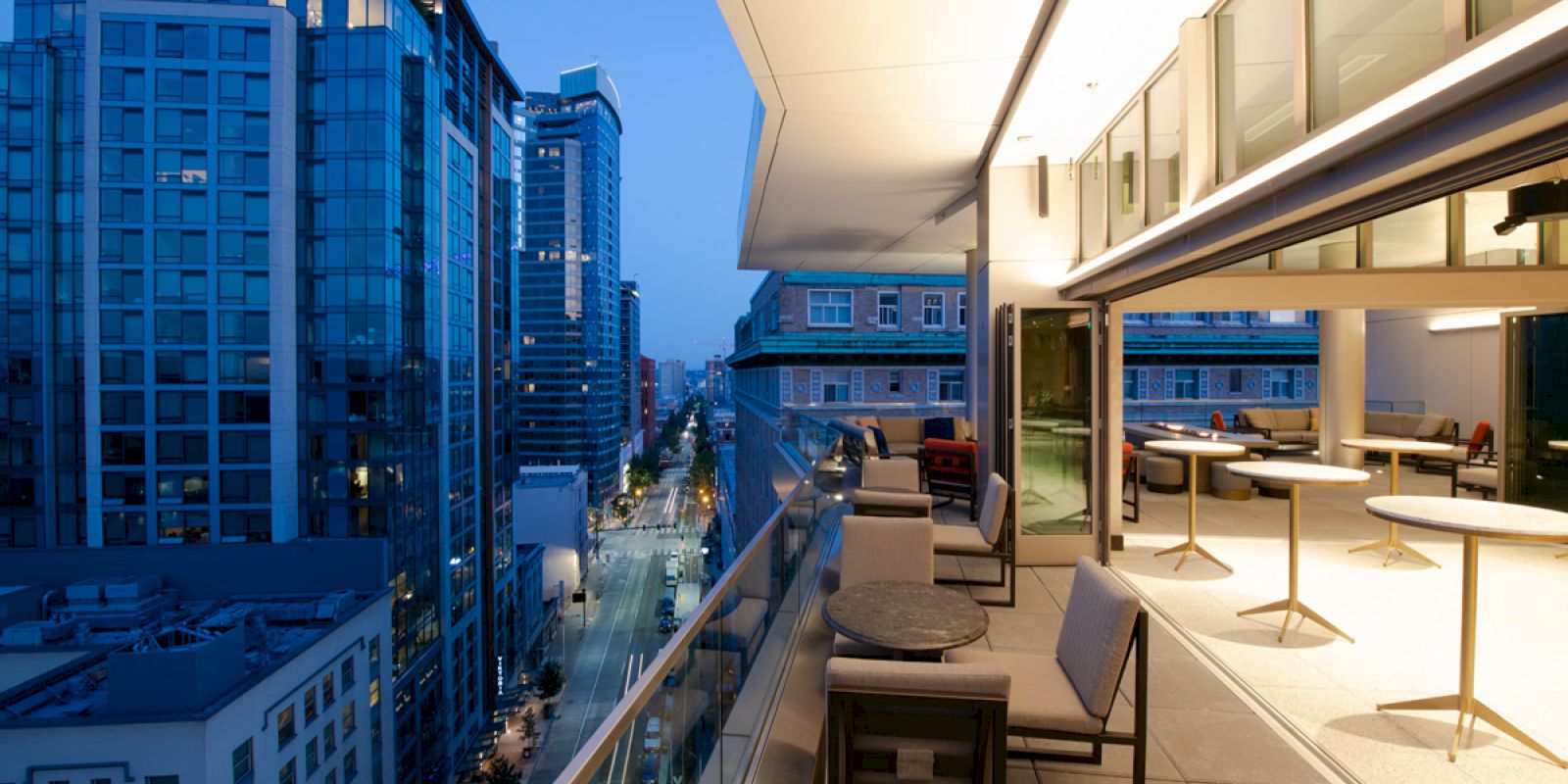 A modern balcony in a high-rise building overlooks a city street at twilight, with outdoor seating and tables.