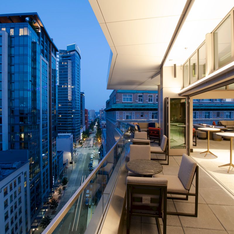 A modern urban balcony view at dusk, featuring outdoor seating, tables, and high-rise buildings lit up alongside a street below.
