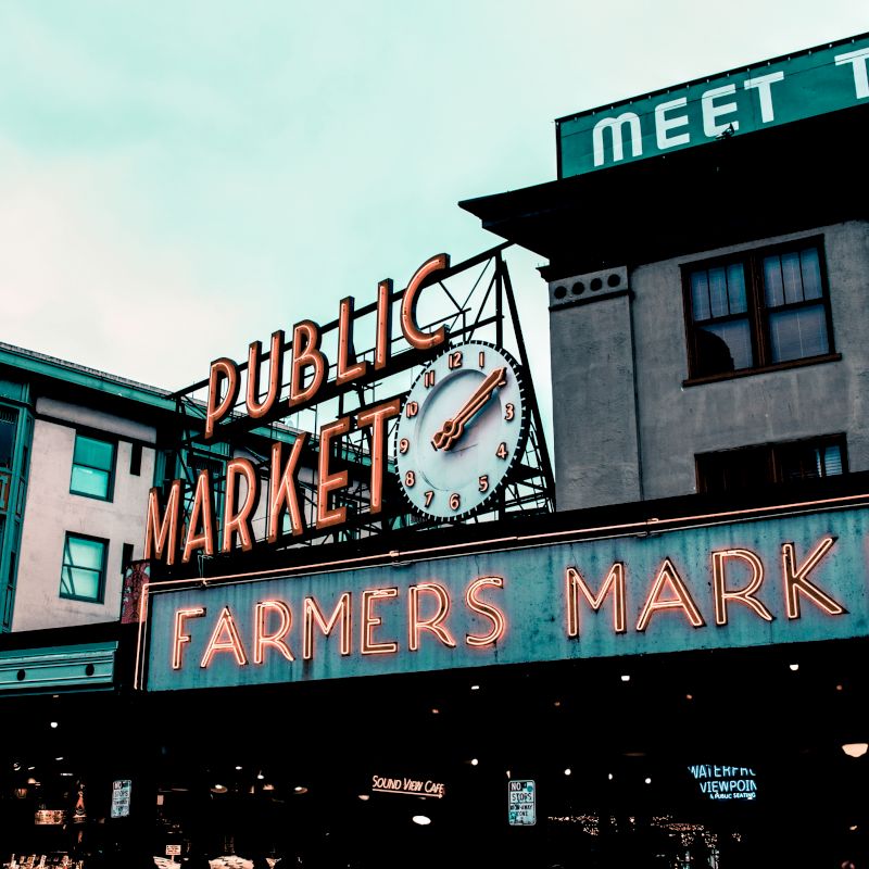 Image shows a neon sign reading "Public Market" and "Farmers Market" attached to a building, likely part of an outdoor market space.