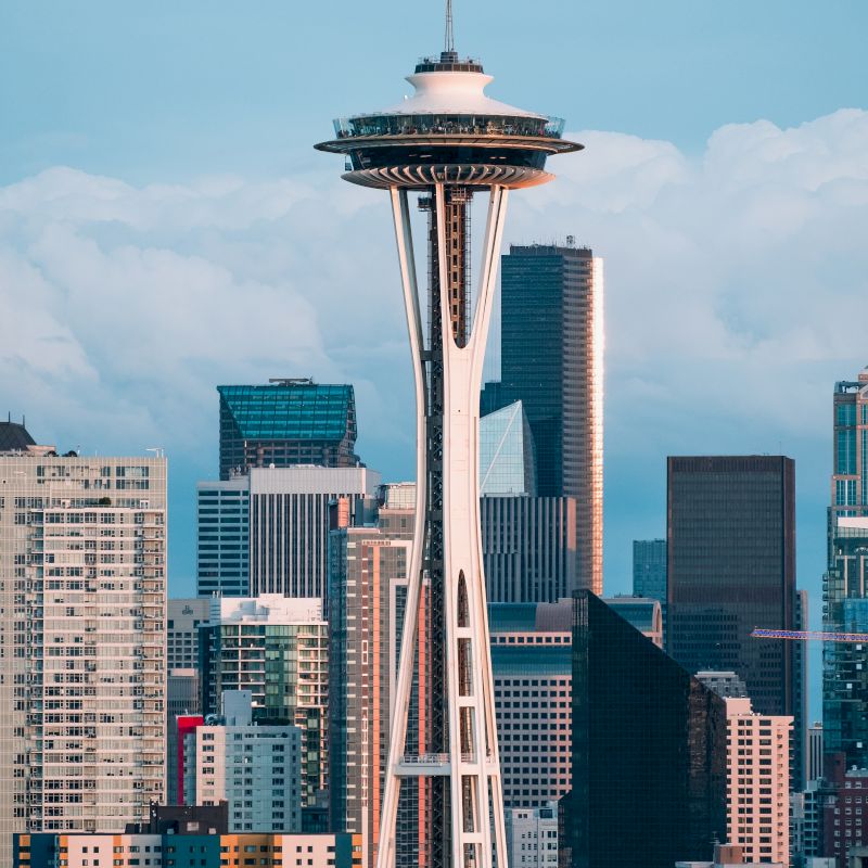 The image shows the Space Needle in Seattle, with the downtown skyline in the background under a partly cloudy sky, highlighting the city's urban landscape.