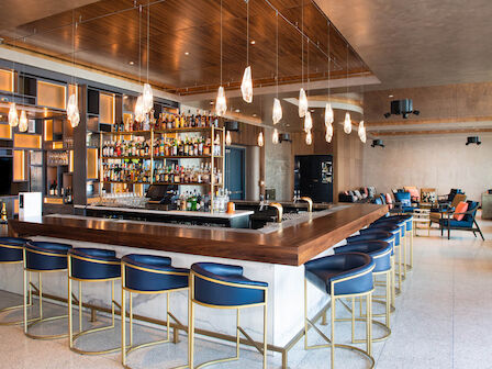 A modern bar with a central counter surrounded by blue bar stools, hanging lights, and a well-stocked shelf of bottles.