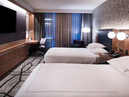 The image shows a modern hotel room with two double beds, a flat-screen TV, a desk by the window, and contemporary decor.