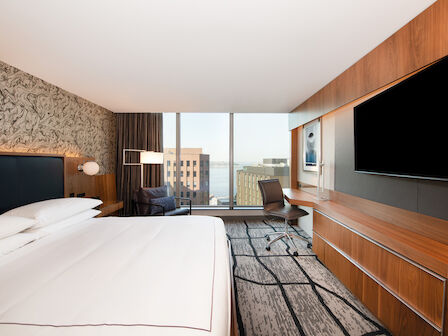 A modern hotel room with a large bed, a desk with a chair, and a sitting area by the window overlooking a cityscape with a partial water view.