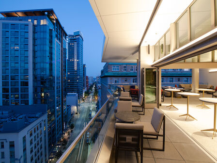 A modern balcony at dusk overlooking a city street with tall buildings. There are tables and chairs on the balcony, and indoor seating visible inside.