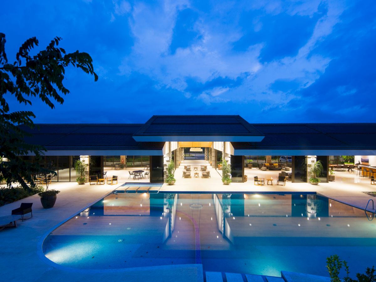 This image shows a large, modern building with a well-lit swimming pool in the foreground at dusk or evening.