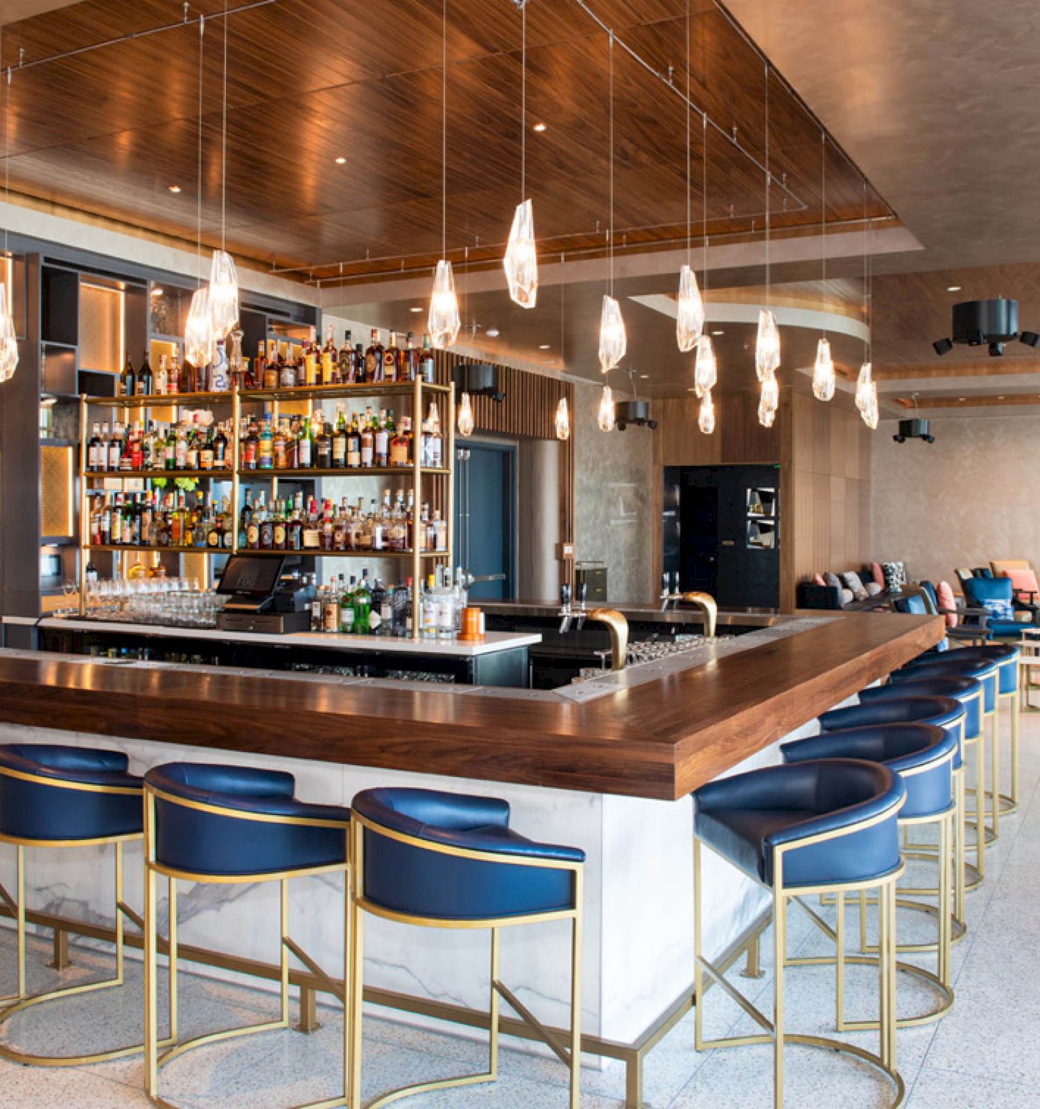 The image shows a modern bar with a wooden counter, blue stools, hanging lights, and a fully stocked liquor shelf in a stylish, spacious room.
