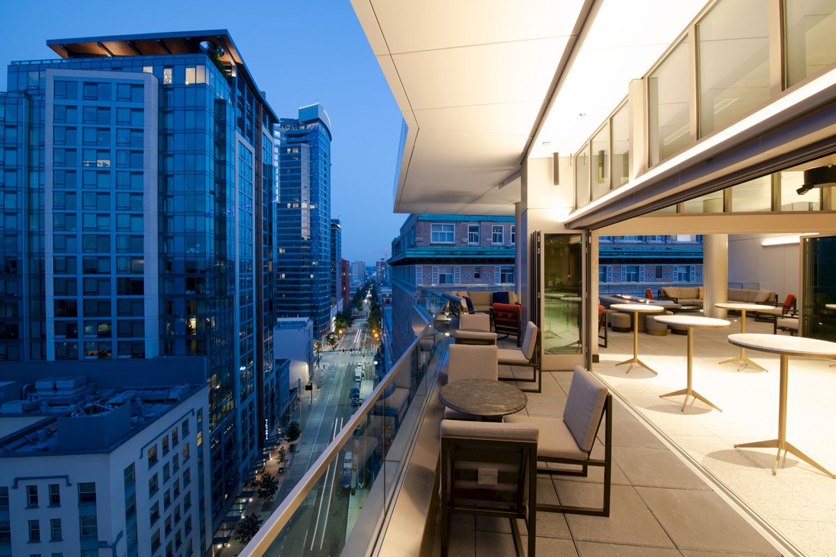 A modern balcony view at dusk with outdoor seating, overlooking a cityscape of tall buildings and a street with lights.