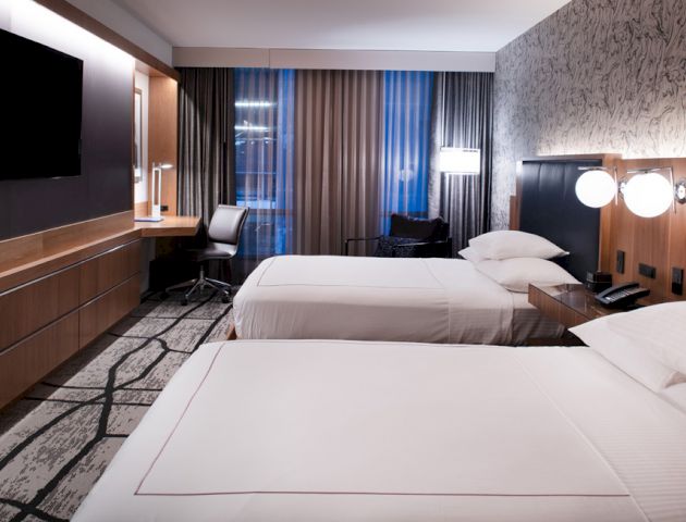 This image shows a modern hotel room with two neatly made beds, a flat-screen TV, a desk with a chair, and stylish decor with ample lighting.