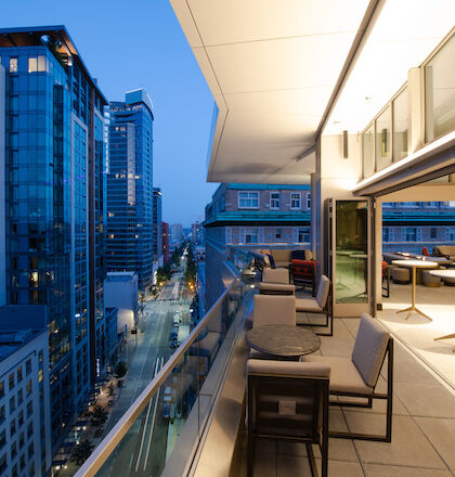 A modern rooftop balcony with seating offers a view of a city street lined with tall buildings during twilight hours.